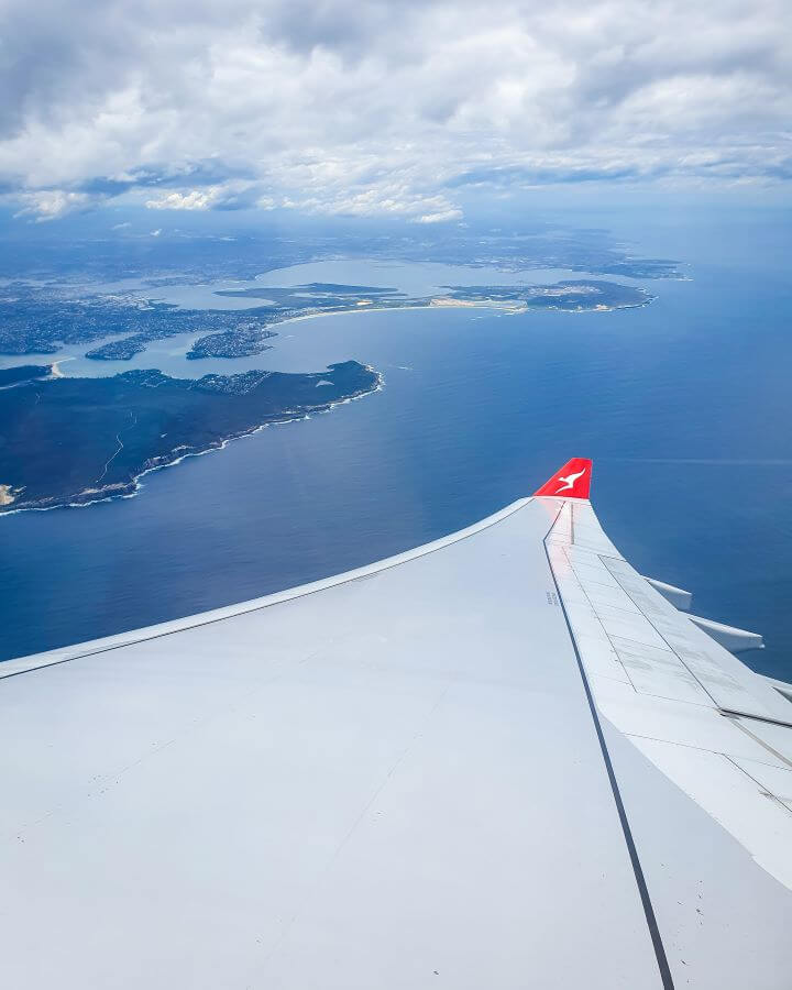 A photo from a plane window down the wing of the plane, which has a red tip with a kangaroo on it, a classic Qantas icon. Beyond the wing is a view of Sydney harbour and the blue water around it "Creating a Realistic Travel Budget: What You Need to Consider"