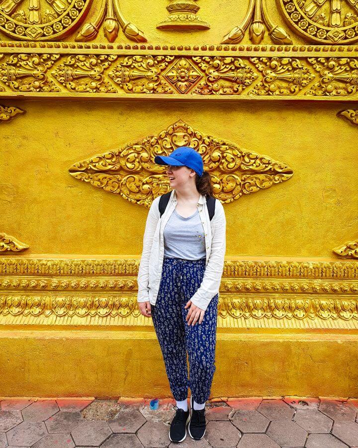 Krissie standing in front of a gold wall with lots of carvings in it. She's wearing long blue pants, a long sleeve top and a blue cap "Creating a Realistic Travel Budget: What You Need to Consider"