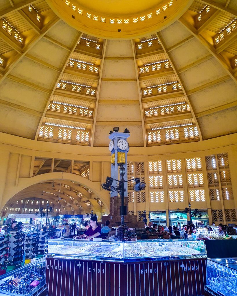 Inside the central market in Phnom Penh, Cambodia. The photo is taken under a large yellow dome with a clock tower and some shop stalls with people standing at them underneath "The Best Six Phnom Penh Activities For Budget Travellers"