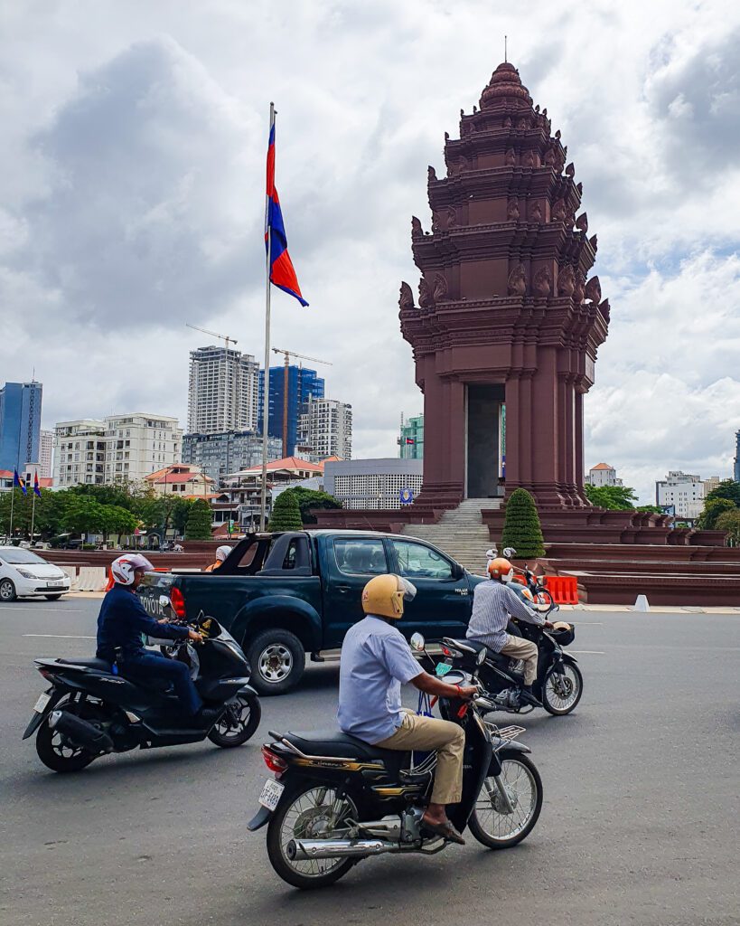 A road with motorbikes and cars on it with a large brown monument behind them in Phnom Penh, Cambodia. The monument is tiered upwards and looks a bit like an upside down acorn on poles. Next to the monument is the Cambodian flag and some other buildings are further behind the monument  "The Best Six Phnom Penh Activities For Budget Travellers"