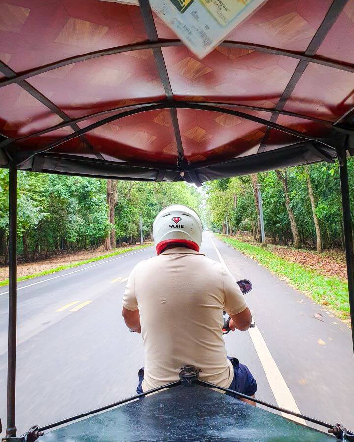 A picture taken while Katherine and Krissie were on the back of a tuk tuk in Siem Reap Cambodia. You can see the roof of the tuk tuk with the driver in a helmet in the middle of the photo. The tuk tuk is on an empty road with lots of bushy trees around "Creating a Realistic Travel Budget: What You Need to Consider"