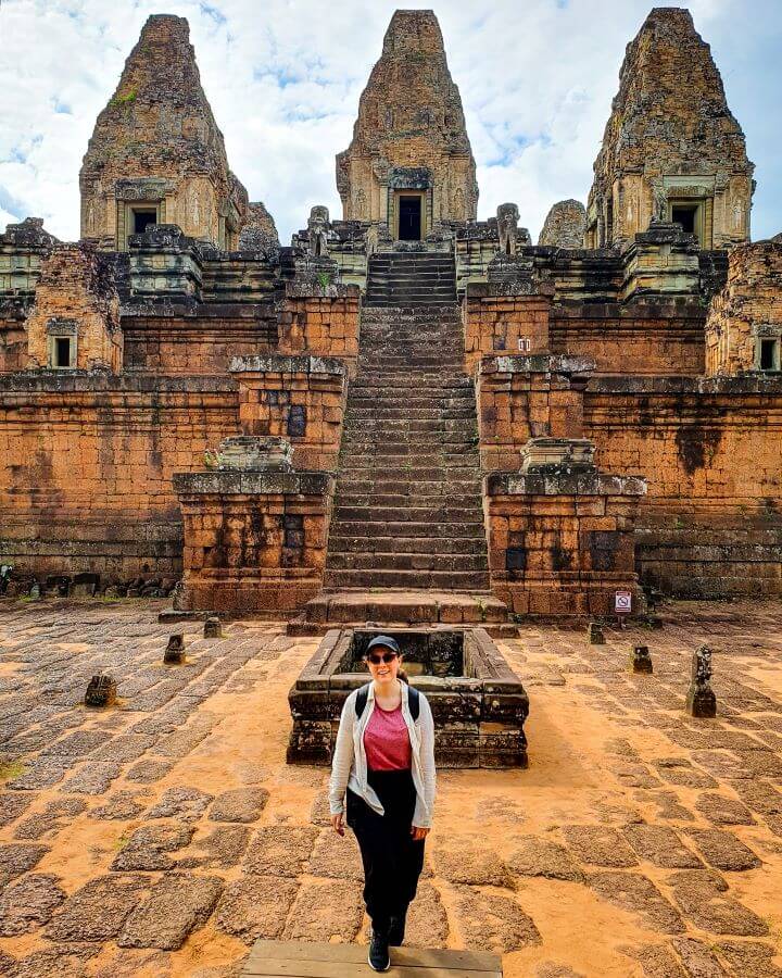 Krissie smiling and standing in front of the very orange and red Pre Rup temple in Siem Reap, Cambodia. The temple has three little towers with doors in them and a huge staircase leading up to them and right behind Krissie is a rectangular brick ledge "Creating a Realistic Travel Budget: What You Need to Consider"