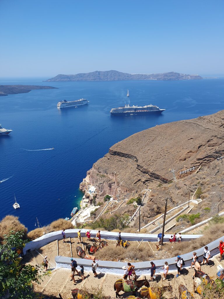 People walking down a long winding staircase that goes down a brown hill in Fira, Santorini, Greece. Behind the hill is the caldera which is a big open space of blue water with more brown land in the distance. You can also see three cruise ships in the water "A Complete Guide To The Santorini Public Transport System"