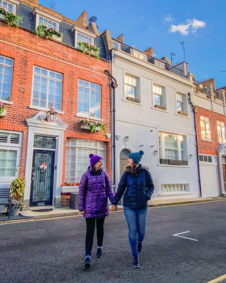 Creating a realistic travel budget. Krissie and Katherine holding hands and walking down a street in London, England. Behind them are some houses made of brick with plants on the windowsills and one house painted grey "Creating a Realistic Travel Budget: What You Need to Consider"