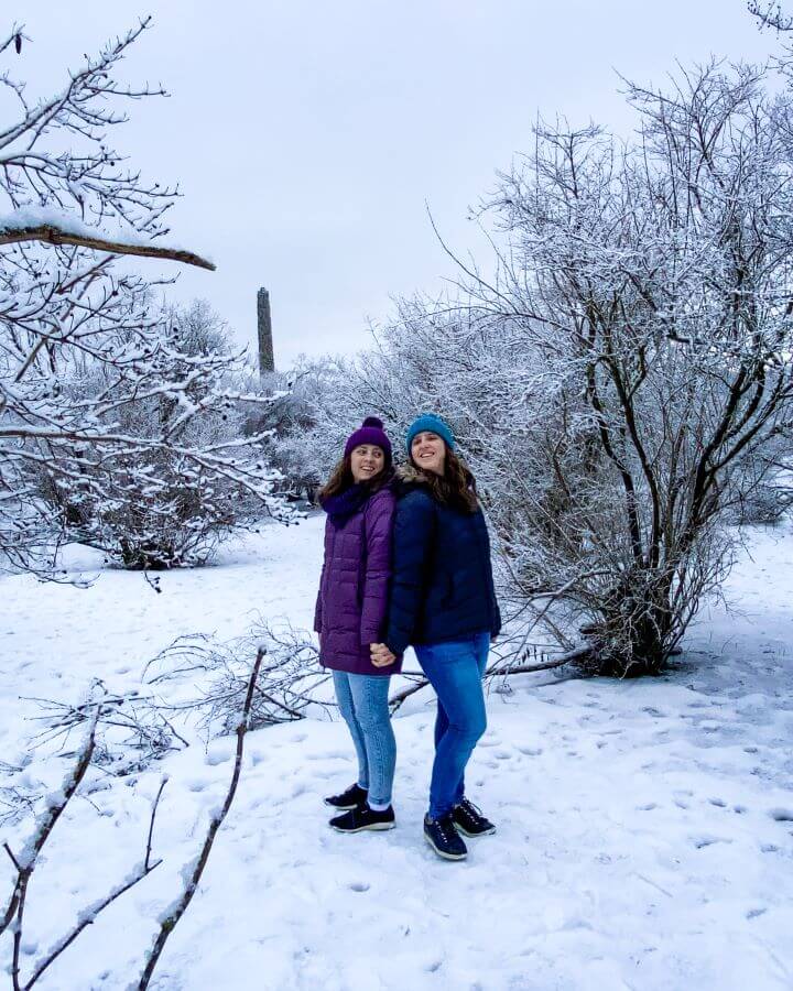 Krissie and Katherine back to back in a snow covered park with snow covered trees all around them. They're wearing puffer jackets and beanies and there's a tall tower in the background "Creating a Realistic Travel Budget: What You Need to Consider"