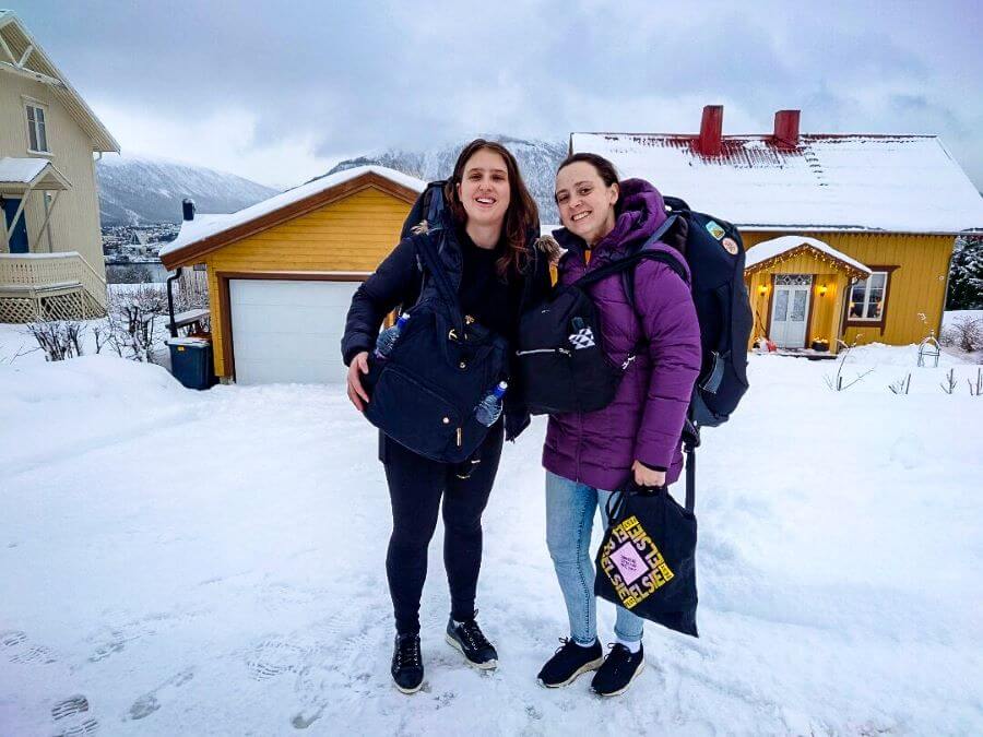 Katherine and Krissie smiling with packs on their backs and front. Behind them is a yellow house and garage and there is snow everywhere! "Creating a Realistic Travel Budget: What You Need to Consider"