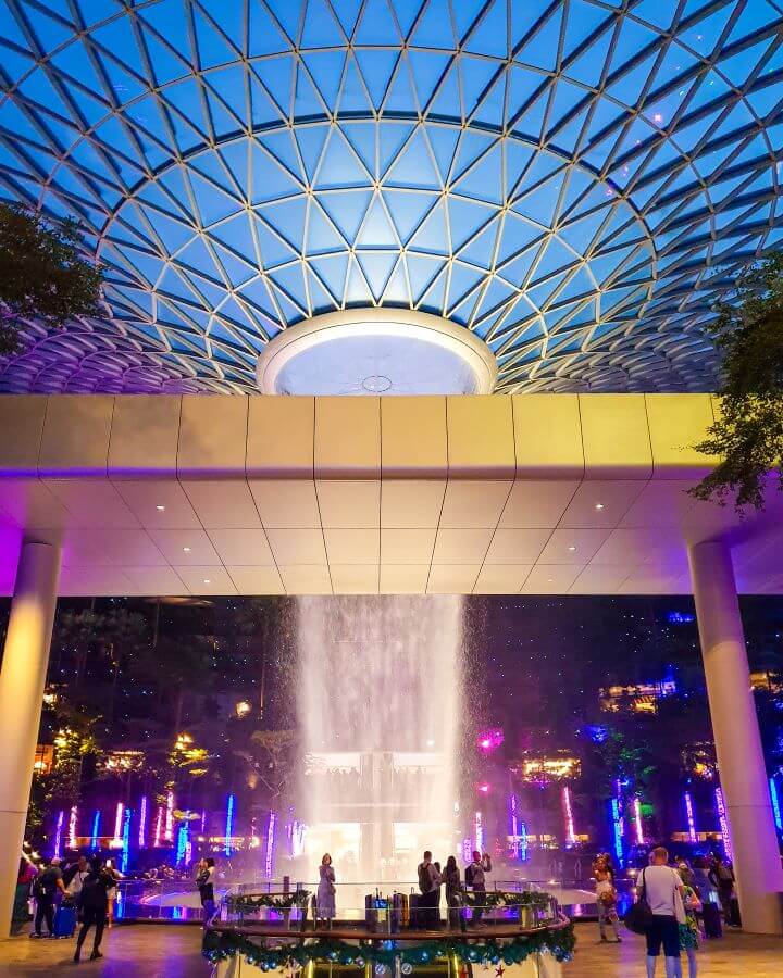 A picture of the giant waterfall coming from the big glass roof at the Jewel in Changi Airport, Singapore. There are blue and purple lights everywhere with people walking all around "10+ Best Affordable Things to do in Singapore"