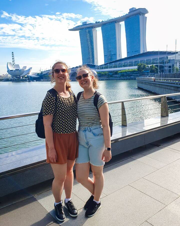 Affordable things to do in Singapore. Katherine and Krissie smiling and standing in front of a metal railing. Behind the railing is the water of the bay in Singapore and the Marina Bay Sands hotel that has 3 skyscrapers with a long platform across the top of all three. In front of the hotel are more buildings including one that looks like a white flower opening "10+ Best Affordable Things to do in Singapore"