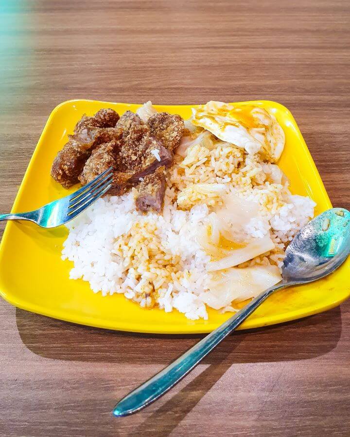 A meal on a yellow plastic plate at a Hawker center in Singapore. On the plate is white rice, crispy pork, some boiled cabbage and a fried egg and it looks very plain but was so tasty! "10+ Best Affordable Things to do in Singapore"