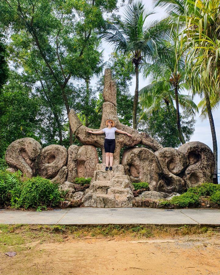 Krissie standing in front of a big sign saying Sentosa on Sentosa Island in Singapore that appears to be made of stone or wood with lots of palm trees behind the sign "10+ Best Affordable Things to do in Singapore"