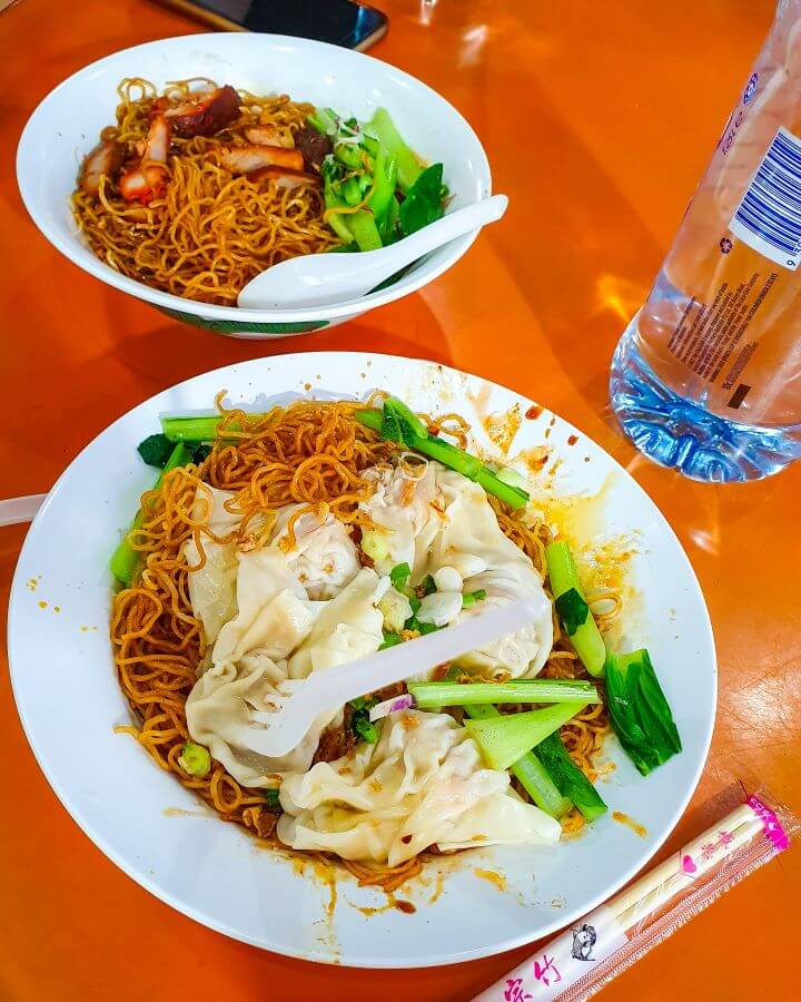 Two bowls of golden coloured noodles with green leafy vegetables in them. One bowl has meat and the other has dumplings in it and they were bought at a Hawker center in Singapore "10+ Best Affordable Things to do in Singapore"