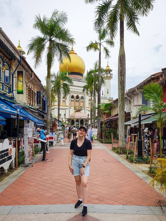 Krissie walking towards the camera on Haji Lane in Singapore. Behind her is a white mosque with a giant gold dome, palm trees and some colourful shops "10+ Best Affordable Things to do in Singapore"