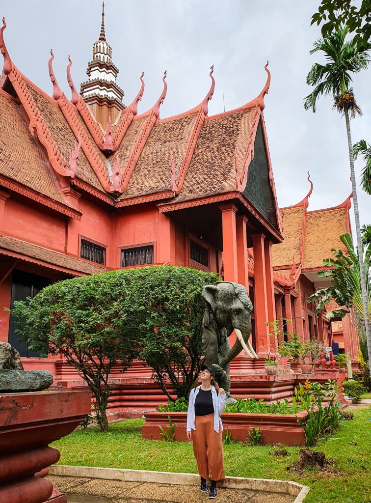 Katherine looking up at the National Museum in Phnom Penh, Cambodia. The museum is pink/orange with a white spire and pointed roof bits. Around the museum are trees and a plant made to look like the body of an elephant, with a little elephant head statue on the front of the bush "The Best Six Phnom Penh Activities For Budget Travellers"