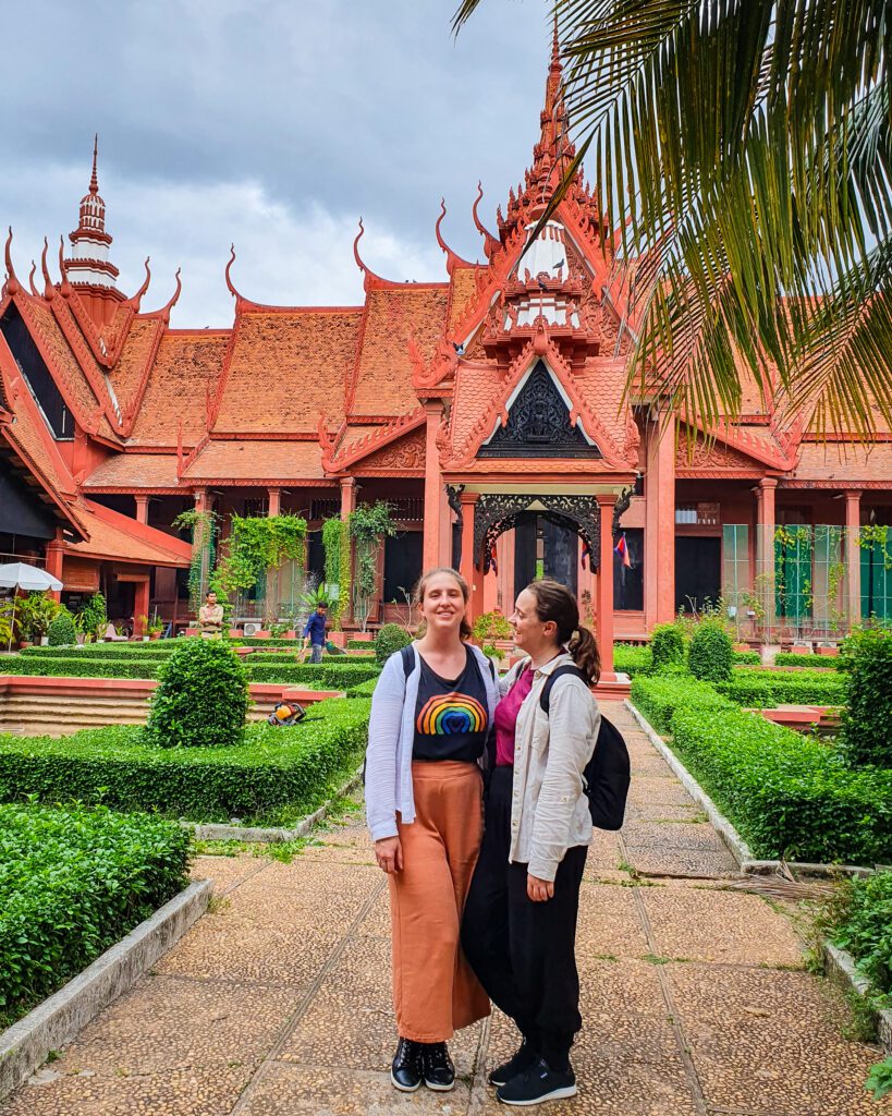 Katherine and Krissie wearing long pants and long sleeve shirts standing on a stone pathway in Phnom Penh, Cambodia. Around the pathway are low green hedges and behind them is a large pink/orange Cambodian style building with spires and lots of pointed roof bits "The Best Six Phnom Penh Activities For Budget Travellers"