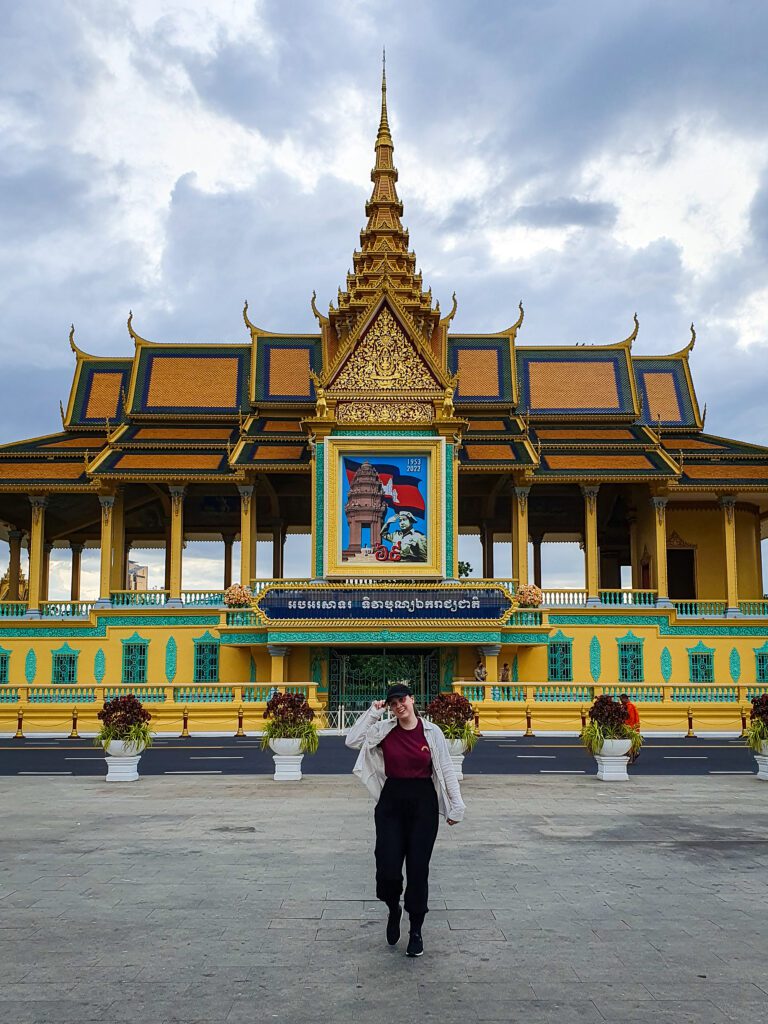 Krissie walking towards the camera in front of the Royal Palace in Phnom Penh, Cambodia. The palace is a large yellow, green, orange and black building with a large golden spire in the middle and pointed roof bits. There are also potted plants and a road out the front "The Best Six Phnom Penh Activities For Budget Travellers"