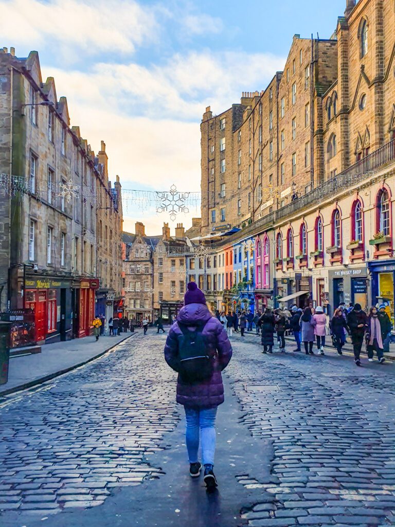 Krissie walking down a stone street in a puffer jacket and beanie in Edinburgh, Scotland. The street has tall brick buildings on both sides with Christmas lights strung up between them. The bottom layer of the buildings are all coloured shops with lots of people walking by "How to See The UK and Ireland Without a Car"