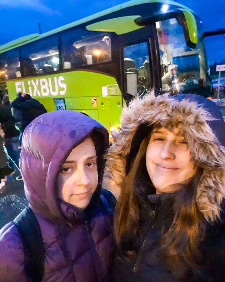 Katherine and Krissie wearing puffer jackets looking very unimpressed in front of a green double decker FlixBus. The light outside shows that it was early morning "FlixBus: The Best Budget Way to Travel Europe Easily"