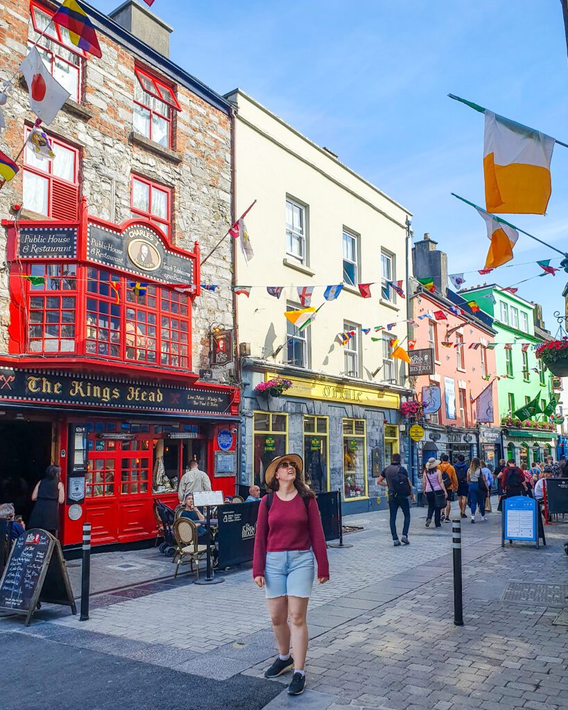 Krissie standing and smiling in the Latin Quarter in Galway, Ireland. Behind her is a colourful street with flags of the world strung between the buildings and lots of pubs and people walking around "How to See The UK and Ireland Without a Car"