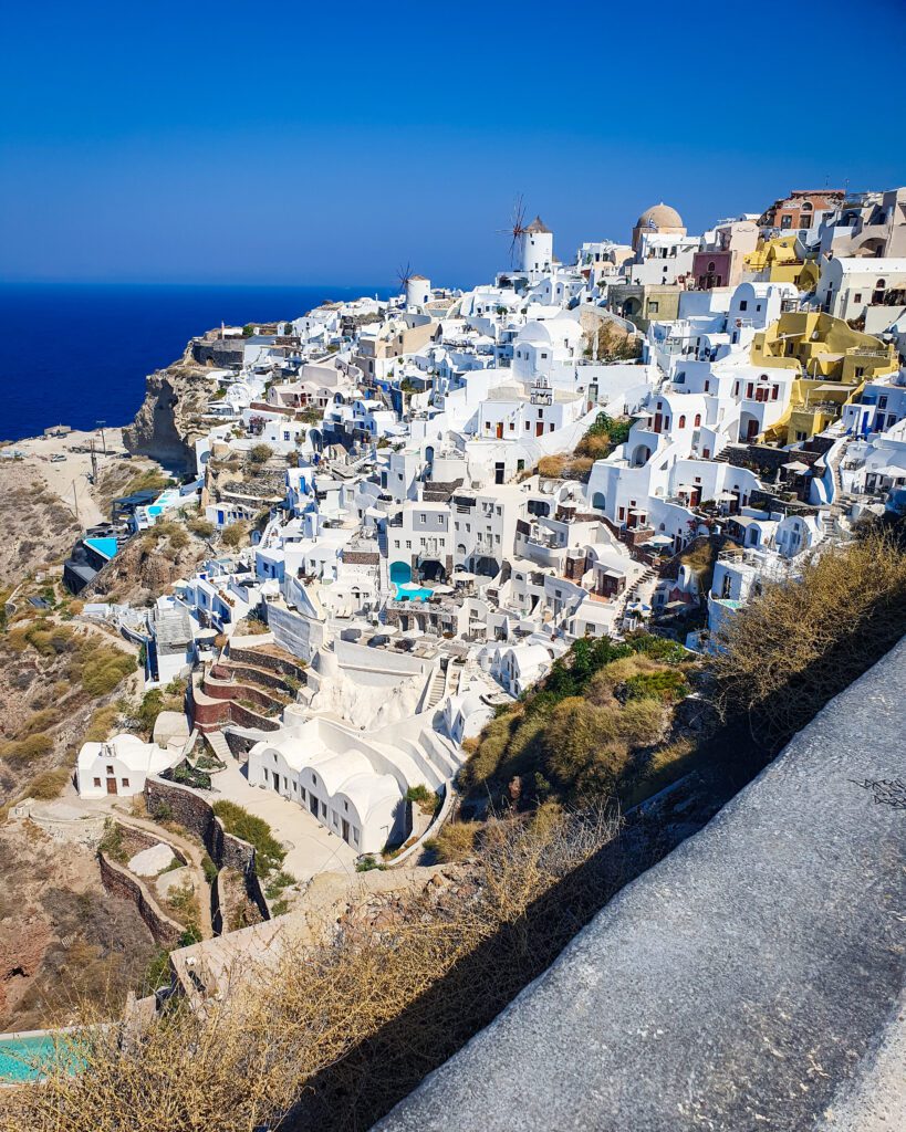 A hill in Santorini, Greece with heaps of white buildings going down the hill. There's a windmill in the distance and some blue water can be seen behind the hill "A Complete Guide To The Santorini Public Transport System"