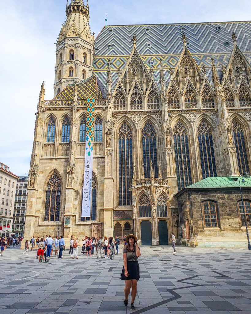 Katherine standing in front of a huge golden cathedral in Vienna, Austria. The cathedral has huge arched windows, pointed parts of the roof while the main roof is tiled in blue, white and yellow tiles "Free and Budget Friendly Ways to Spend Your Time in Vienna"