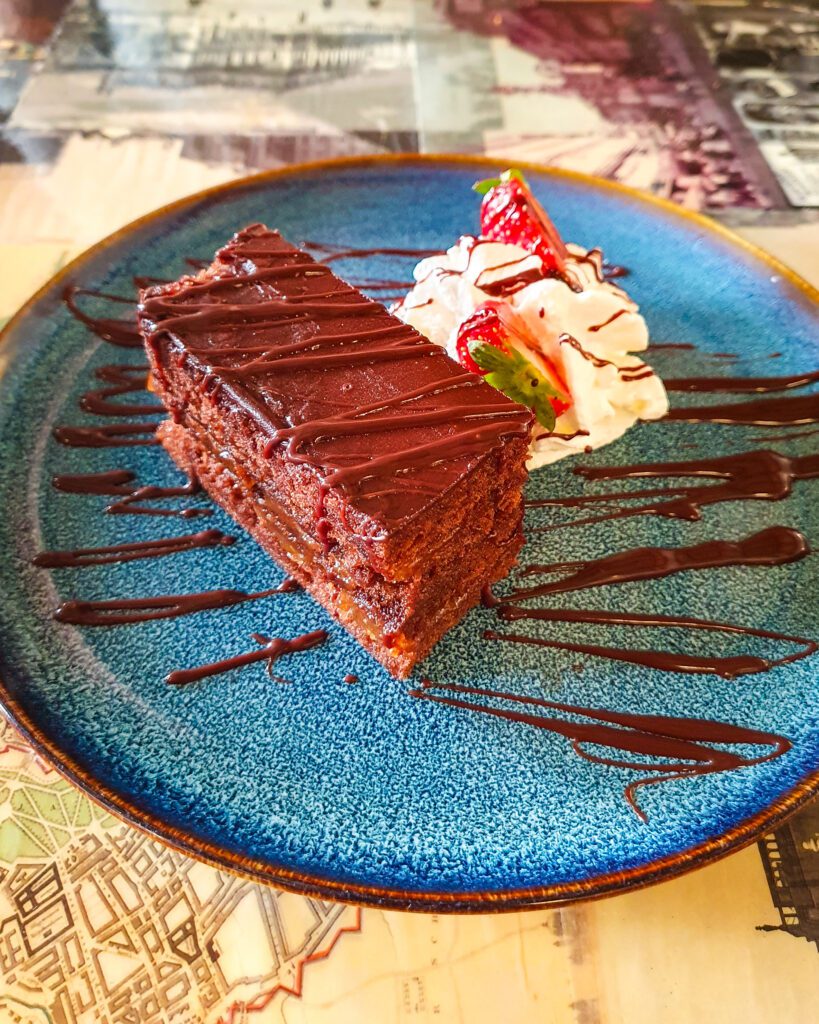 A blue plate with a rectangular slice of sachertorte in Vienna. Austria. The cake is a chocolate cake with a chocolate drizzle over the top with a blob of whipped cream and strawberries next to it "Free and Budget Friendly Ways to Spend Your Time in Vienna"