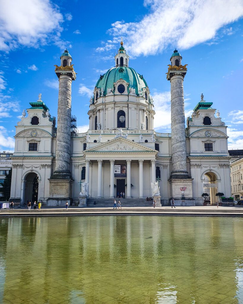 Across a yellow pond is a church with 2 large round towers and a huge blue dome in the middle in Vienna, Austria "Free and Budget Friendly Ways to Spend Your Time in Vienna"