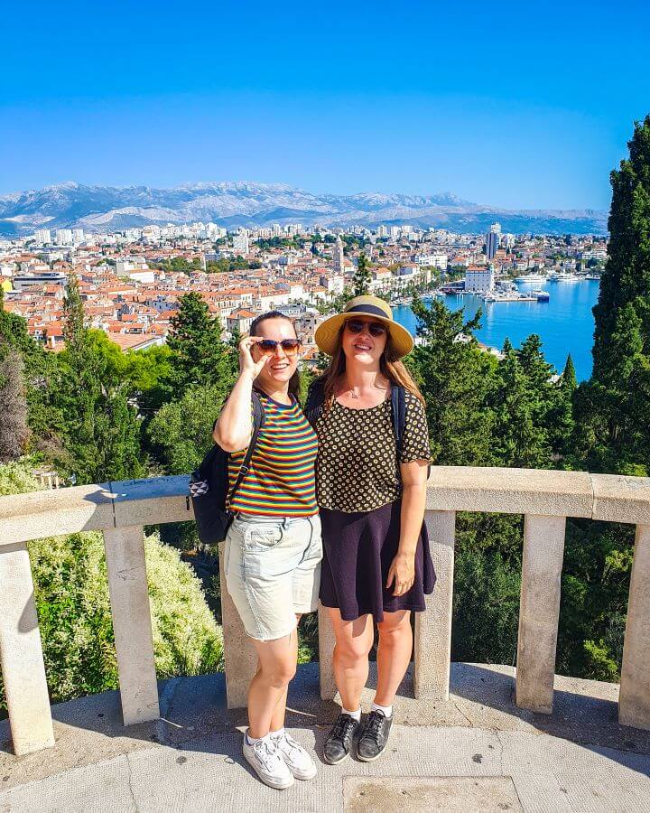 Krissie and Katherine smiling and standing in front of a barrier with lots of green trees and a view of Split, Croatia behind them. The city with its orange roofs is on the left and the blue water is on the right. Behind the city are big hills "FlixBus: The Best Budget Way to Travel Europe Easily"