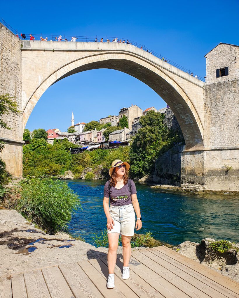 Krissie smiling and standing in front of the Stari Most bridge in Mostar, Bosnia and Herzegovina. It is a tall but small bridge that arches that sharply up in the middle with people on top of the bridge and a blue river underneath "How To Decide On The Better Day Trip From Dubrovnik"