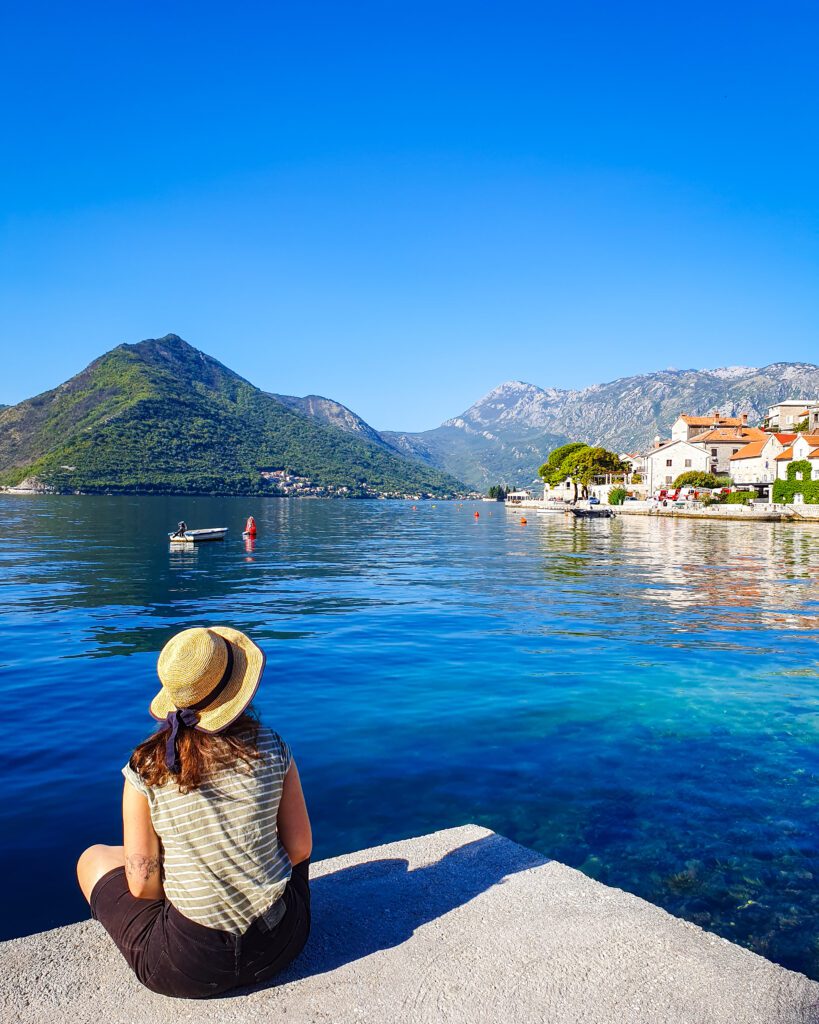 The best day trip from dubrovnik. Krissie sitting in front of a large body of water which is Kotor Bay in Montenegro. Behind her are some huge mountains and a little town "How To Decide On The Better Day Trip From Dubrovnik"