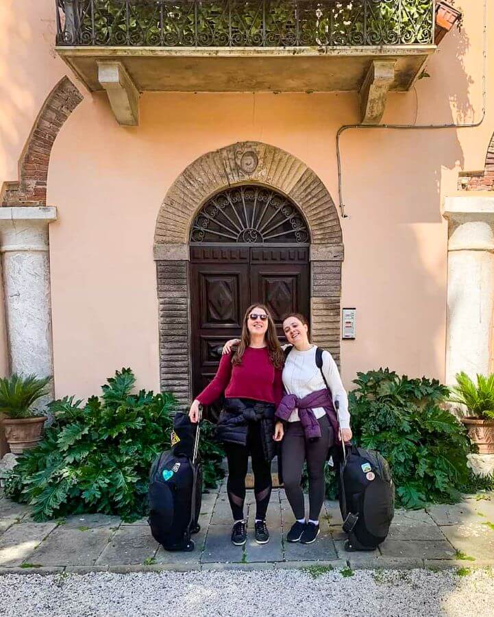 Katherine and Krissie smiling and standing outside a Tuscan villa with one roller suitcase each and puffer jackets tied around their waists "FlixBus: The Best Budget Way to Travel Europe Easily"