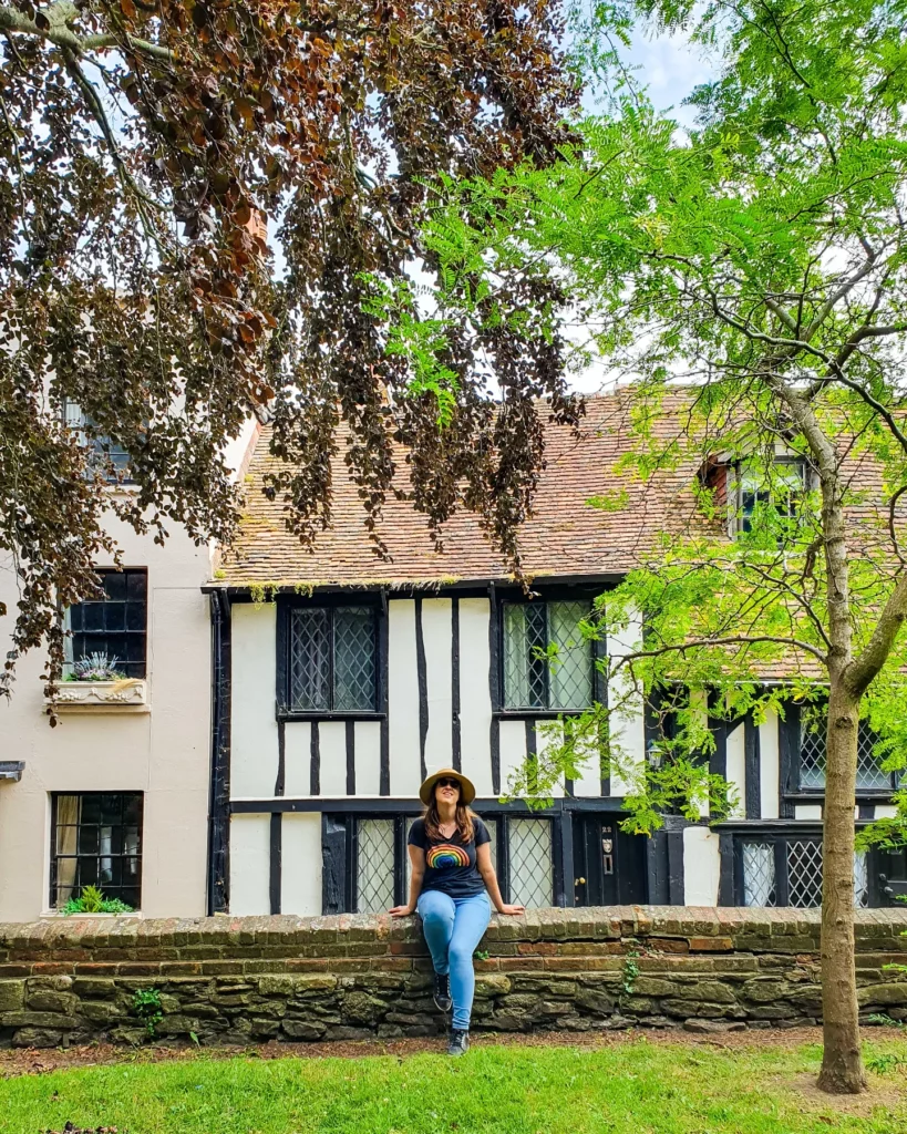 UK and Ireland without a car. Katherine sitting on a stone wall with white old English buildings behind her in Rye, England. The buildings have tiled roofs and black planks of wood vertically through them and there are also some green trees in front of them "How to See The UK and Ireland Without a Car"