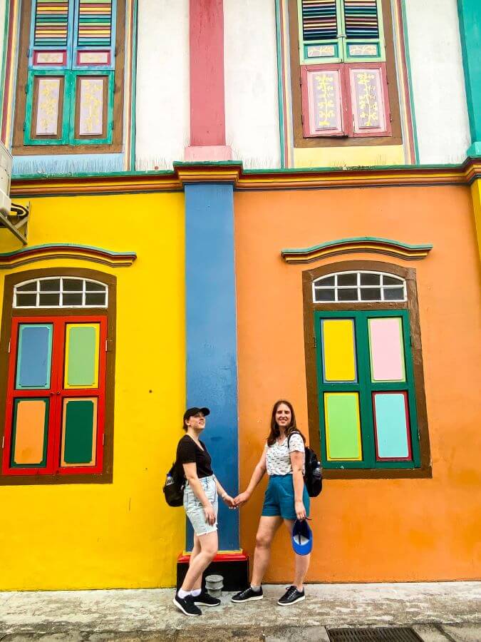 Krissie and Katherine holding hands and standing in front of the Tan Teng Niah house in Singapore. The house is a really colourful house, behind them is a orange, blue and yellow wall with windows split into quarters and each quarter is a different colour "10+ Best Affordable Things to do in Singapore"