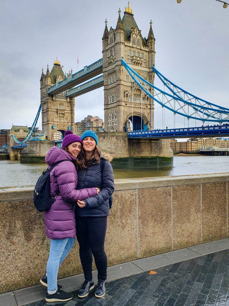 Krissie and Katherine hugging and smiling with puffer jackets and beanies on. Behind them is tower bridge in London, England which has 2 giant stone towers with blue metal strung between them "How to Have the Best Time in London (For Free!)"