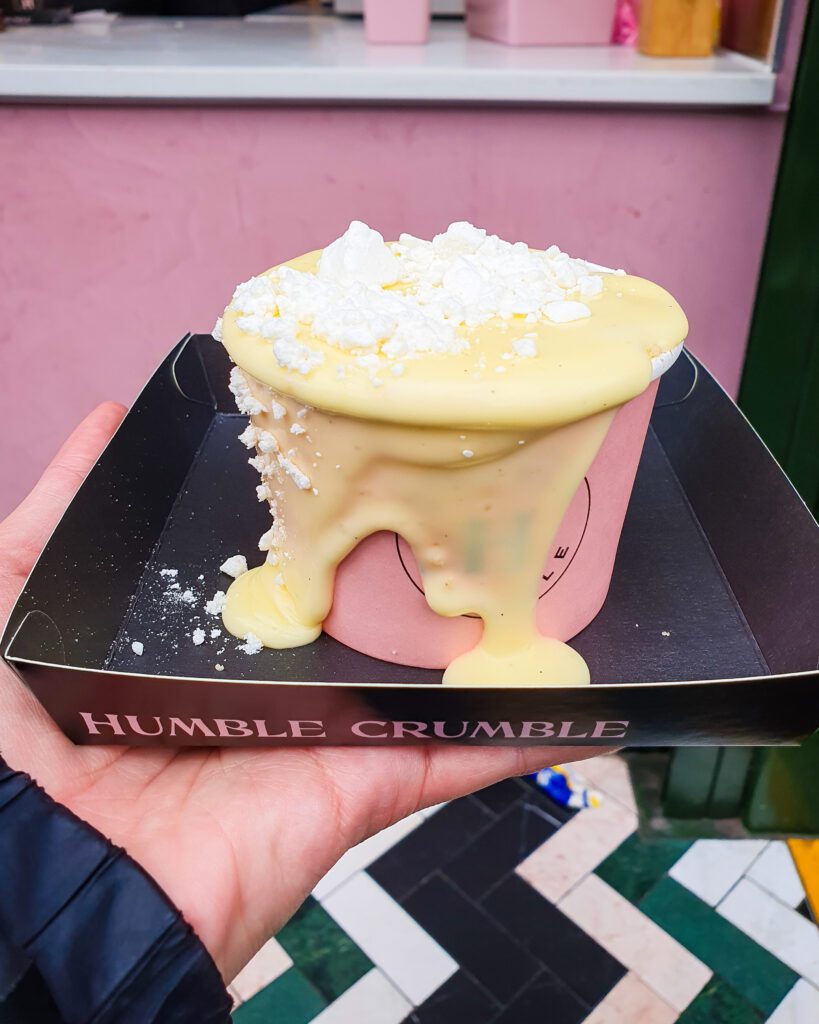 A hand holding a black box that says "Humble Crumble" which is a great dessert place in London, England. In the box is a pink round container overflowing with custard with white meringue on top "How to Have the Best Time in London (For Free!)"