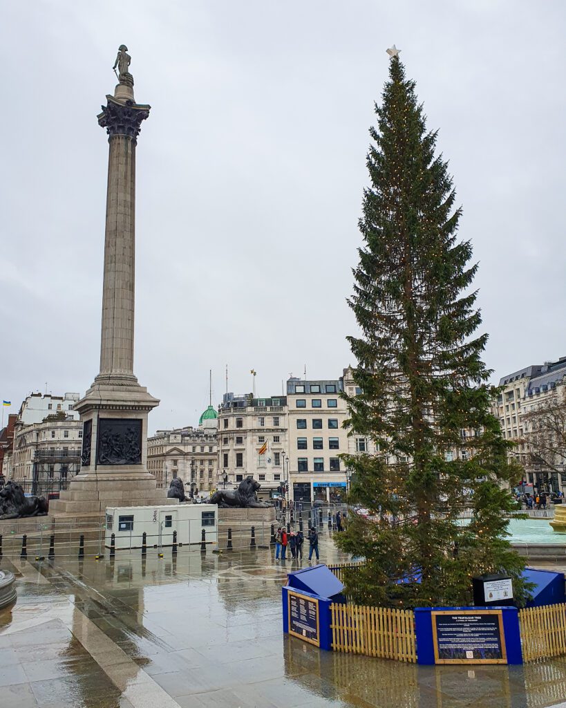 Trafalgar Square in London, England. On the left is a big stone pillar with a stone figure on top and on the right is a large Christmas tree that has very sparse branches "How to Have the Best Time in London (For Free!)"