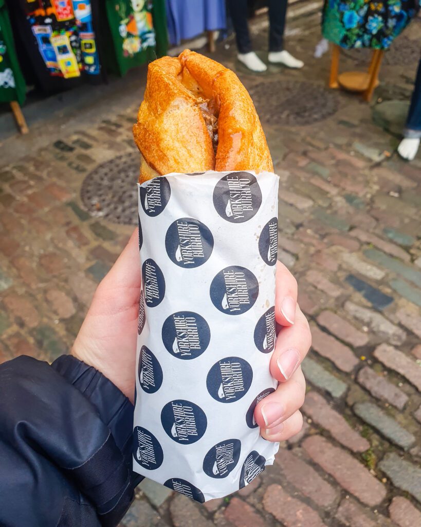 A hand holding a big yorkshire pudding folded up into a burrito and wrapped in paper that says "Yorkshire Burrito" on it which is a great place in London, England "How to Have the Best Time in London (For Free!)"