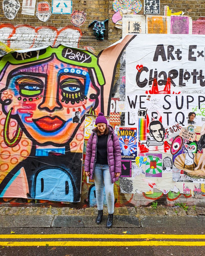 Krissie standing in front of some colourful street art and posters in Brick Lane, London, England "How to Have the Best Time in London (For Free!)"