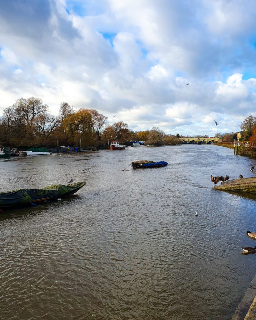 The Thames river in Richmond, England. There's boats on the river and trees with a bridge in the distance "How to Have the Best Time in London (For Free!)"