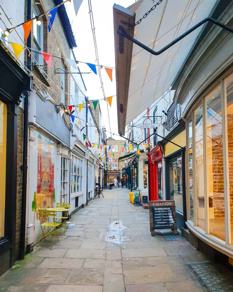 A small street in Richmond, England. There's shops on either side of the stone street and flags strung up above the shops "How to Have the Best Time in London (For Free!)"