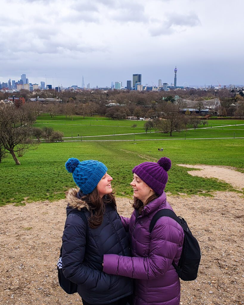 Katherine and Krissie looking at each other and smiling with a big green hill behind them and a view of the London Skyline behind that "How to Have the Best Time in London (For Free!)"