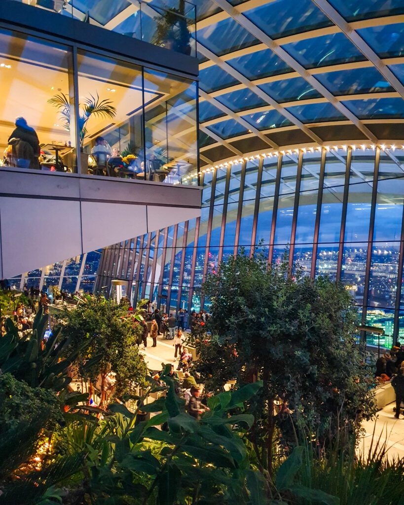 Inside the Sky Garden in London, England which is a big glass building with trees and other plants inside and lots of people walking around "How to Have the Best Time in London (For Free!)"