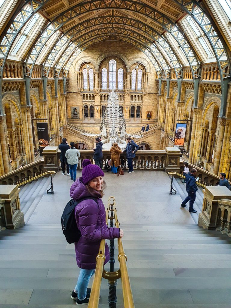 Krissie smiling and standing on a staircase in the Natural History Museum in London, England. Behind her is the big stone main hallway with a huge whale skeleton hanging from the roof and people walking around "How to Have the Best Time in London (For Free!)"