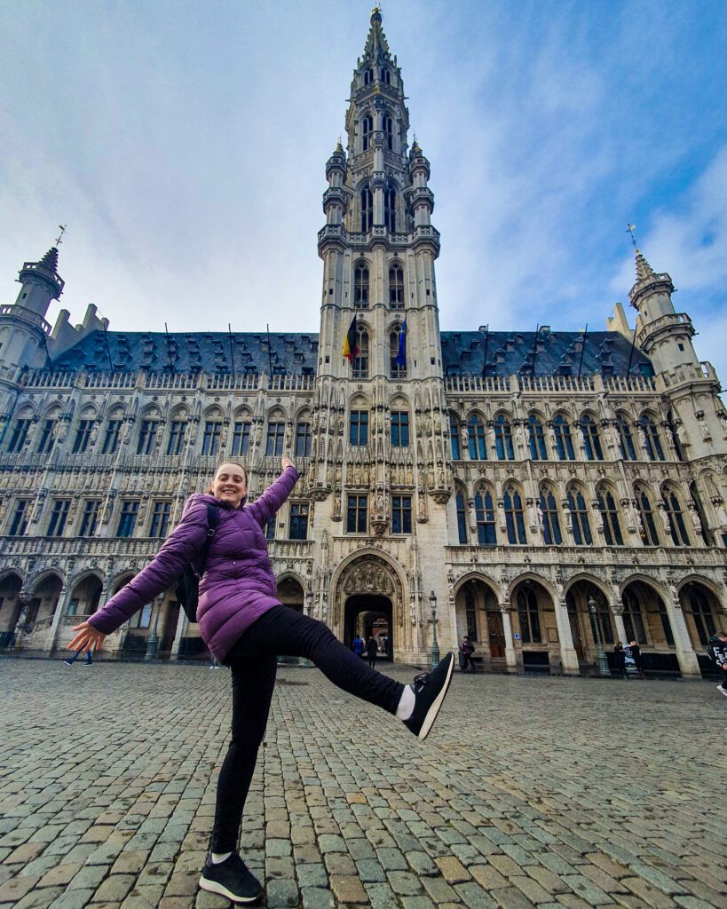 Krissie posing in front of the Brussels Town Hall in Belgium. The Town Hall is a big stone building with a blue roof and has a huge bell tower in the middle "Wonderful Ways to Spend Your Time in Bruges"