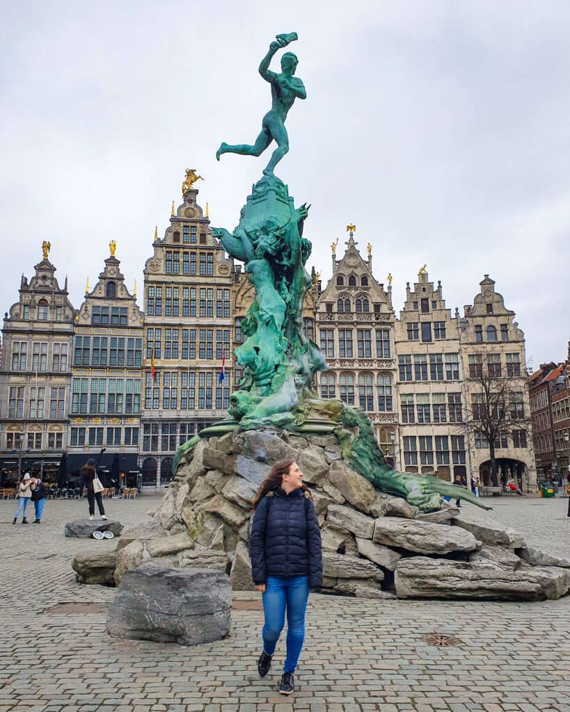 Katherine standing in a town square in Antwerp, Belgium in front of a statue on top of a huge pile of stones with a big green man throwing something on top. Behind the statue are six tall extravagant buildings with gold statues on top of 5 of them "Wonderful Ways to Spend Your Time in Bruges"