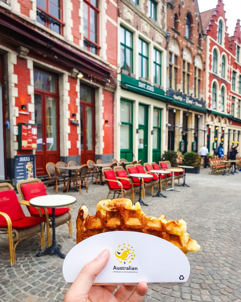 A waffle with a bite out of it with some beautiful coloured buildings behind it in Bruges, Belgium "Wonderful Ways to Spend Your Time in Bruges"