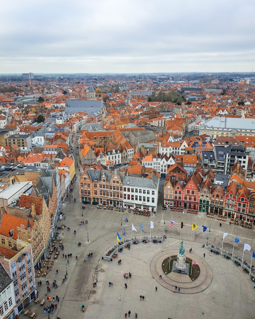 A view down at Grote Markt from the Belfry Tower in Bruges, Belgium. The bottom third of the view is the square with people walking around and a statue in the middle. The rest of the view is lots of buildings with pointed orange roofs "Wonderful Ways to Spend Your Time in Bruges"