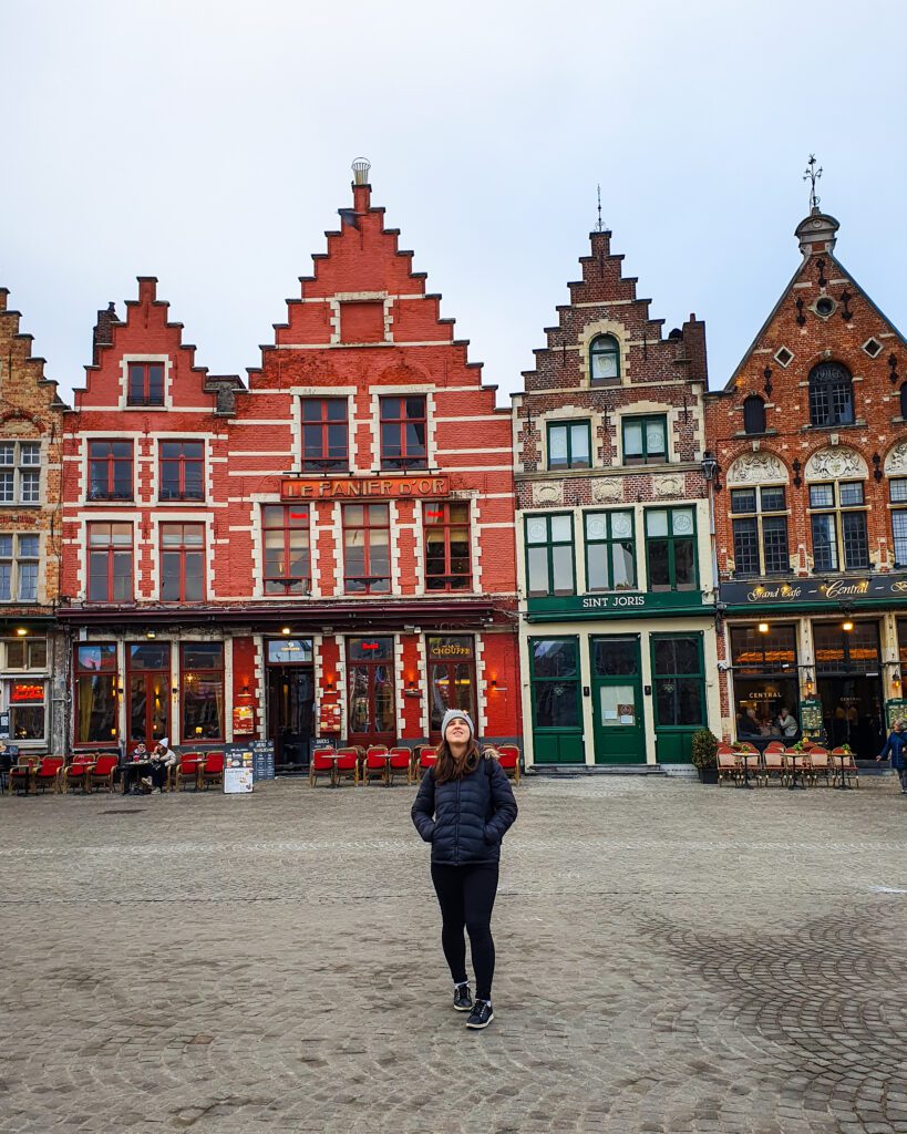 Katherine standing and looking up in a town square called Grote Markt in Bruges, Belgium. Behind her are 4 coloured buildings with stepped roofs to a triangular point "Wonderful Ways to Spend Your Time in Bruges"