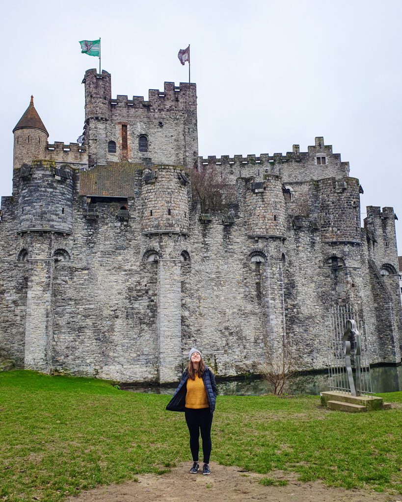 Katherine standing on some grass in front of the Gravensteen castle in Ghent, Belgium. The castle is made of grey stone and has lots of turrets and 2 flags on the top "Wonderful Ways to Spend Your Time in Bruges"