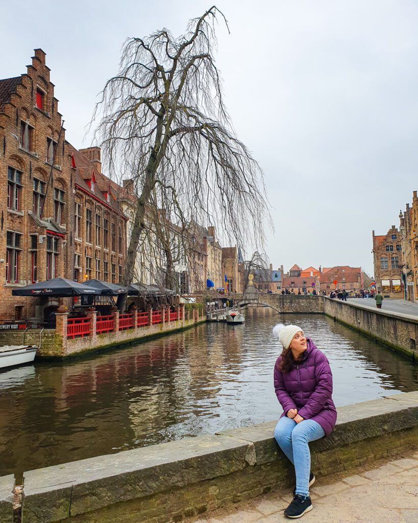 Krissie sitting on the side of the Bruges canal. There are lots of beautiful orangey red buildings and some trees behind her as well as the canal and another bridge in the background "Wonderful Ways to Spend Your Time in Bruges"