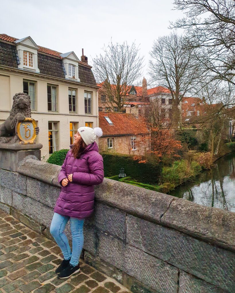 Krissie leaning against a stone barrier of a bridge over some water in Bruges, Belgium. Beyond the bridge are some houses, bare trees and other green plants "Wonderful Ways to Spend Your Time in Bruges"